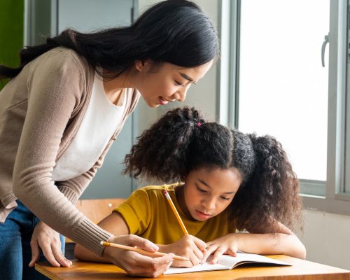 Asian school teacher assisting female student in classroom. Young woman working in school helping girl with her writing, education, support, care.